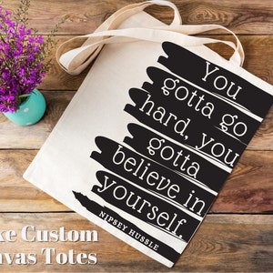 K&ouml;nnte beinhalten: Eine wei&szlig;e Canvas-Tragetasche mit schwarzen, gestrichenen Streifen und dem Text "You gotta go hard, you gotta believe in yourself." Der Text wird "NIPSEY HUSSLE" zugeschrieben.