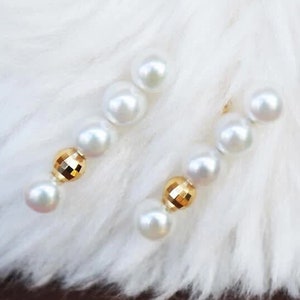 May include: A pair of gold earrings with white pearls and gold accents. The earrings are on a white fluffy surface. The text "Pearlygummi" is visible in the bottom right corner of the image.