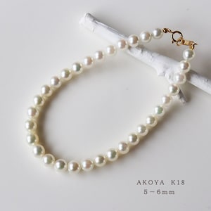 Pode incluir: Um colar de pérolas brancas com um fecho de ouro. As pérolas medem 5-6 mm. O colar está sobre uma superfície de madeira branca. AKOY A K18 5-6mm