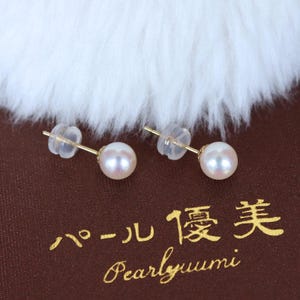 May include: A pair of gold stud earrings with round, lustrous white pearls. The earrings are set against a brown background with Japanese text and the English word "Pearlyuumi". The pearls have a soft, pinkish hue.