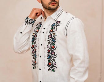 May include: White long-sleeve shirt with colorful floral embroidery down the front and on the cuffs. The shirt has black buttons and decorative stitching. A traditional style garment.
