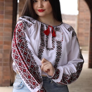 May include: A woman wearing a white embroidered blouse with red and black geometric patterns. The blouse has a red tassel at the neckline and a small tag with the text "LUKART" on the right side. She is wearing blue jeans.