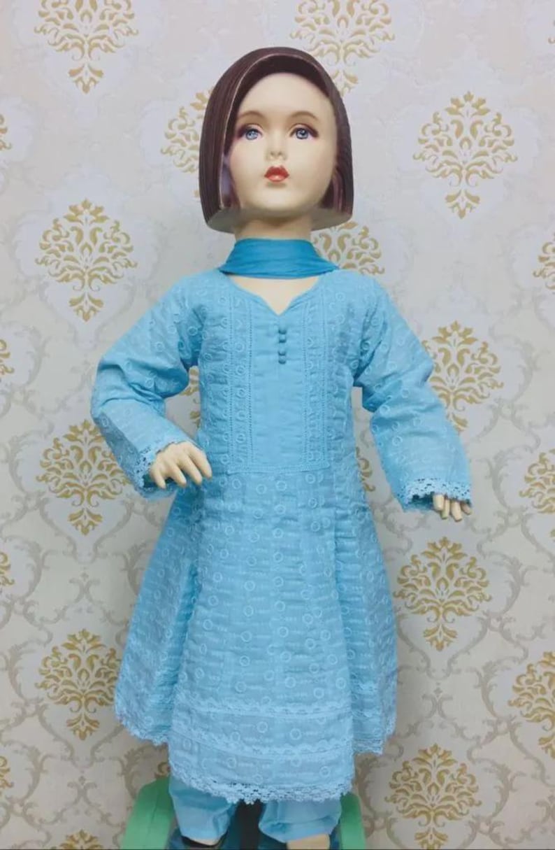 May include: A light blue, long-sleeved dress with intricate embroidery and lace trim, paired with matching pants and a scarf. The dress features a V-neck and button details. The outfit is displayed on a mannequin.