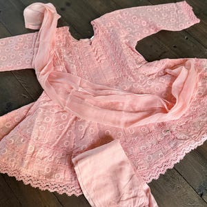 May include: A light pink, embroidered dress set with matching pants and a head covering. The dress features a round neckline, long sleeves, and a scalloped hem. The fabric has a delicate, eyelet-like pattern.