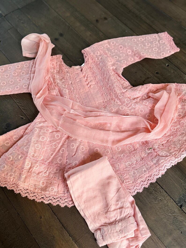 May include: A light pink, embroidered dress set with matching pants and a head covering. The dress features a round neckline, long sleeves, and a scalloped hem. The fabric has a delicate, eyelet-like pattern.