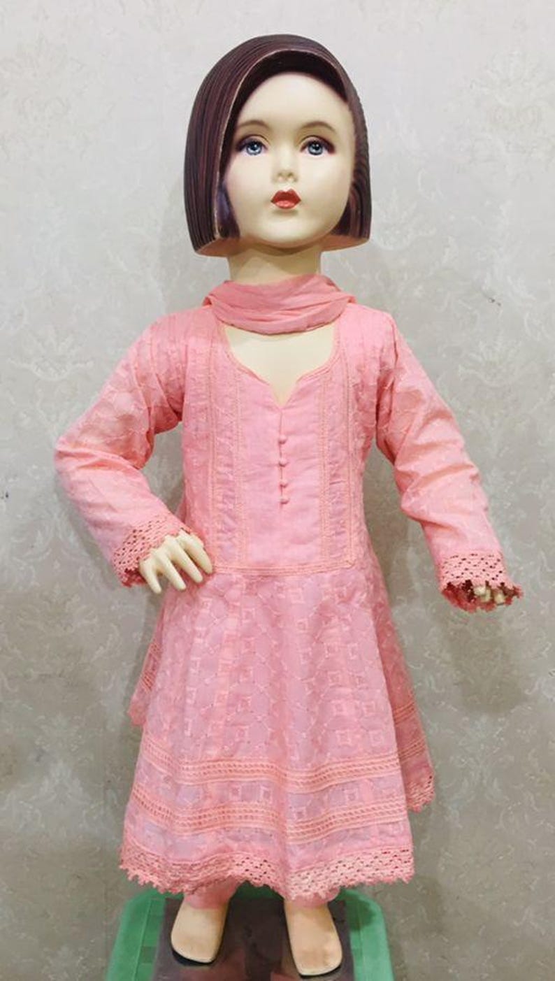 May include: A pink embroidered dress with long sleeves and a matching scarf, displayed on a mannequin. The dress features intricate lace detailing on the sleeves and hem. The mannequin has a brown wig and is standing on a green surface.