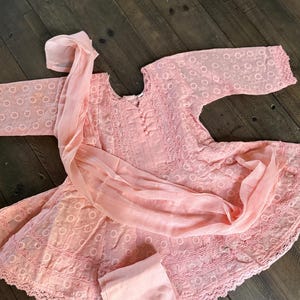 May include: A light pink embroidered outfit, including a tunic, pants, and a scarf. The tunic features a round neckline and long sleeves. The fabric has a delicate, openwork design. The outfit is laid out on a dark wood surface.