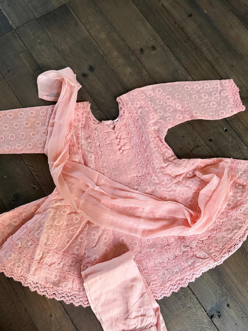 May include: A light pink embroidered outfit, including a tunic, pants, and a scarf. The tunic features a round neckline and long sleeves. The fabric has a delicate, openwork design. The outfit is laid out on a dark wood surface.