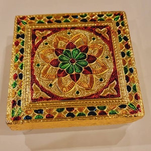 May include: A square, gold-colored box with a decorative, multi-colored design. The design features a central flower with red, green, and yellow petals, surrounded by a geometric pattern.