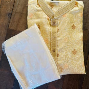 Pu&ograve; includere: Un completo kurta pajama giallo chiaro. Il kurta presenta un colletto alla coreana e finiture dorate decorative. Il tessuto ha un sottile motivo floreale con accenti dorati e pesca. I pantaloni del pigiama sono piegati e avvolti in plastica trasparente.