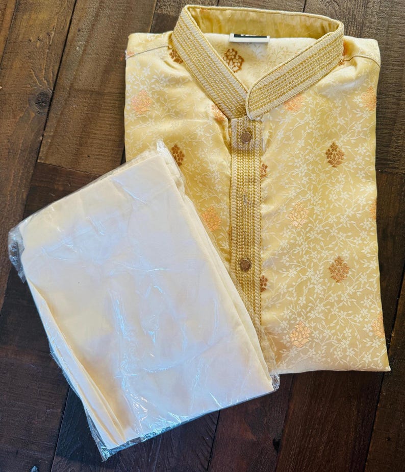 Pu&ograve; includere: Un completo kurta pajama giallo chiaro. Il kurta presenta un colletto alla coreana e finiture dorate decorative. Il tessuto ha un sottile motivo floreale con accenti dorati e pesca. I pantaloni del pigiama sono piegati e avvolti in plastica trasparente.