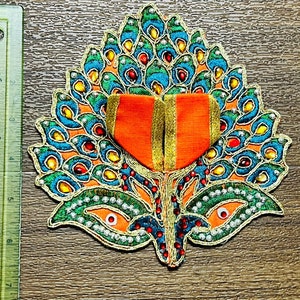 May include: A decorative, embroidered peacock feather design with vibrant colors. The design features teal, orange, and green hues, accented with red and yellow jewels. The center has an orange heart shape with gold trim.