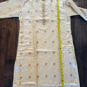 Pu&ograve; includere: Kurta color crema con colletto alla coreana e maniche lunghe. Il tessuto presenta un motivo ripetuto di piccoli motivi floreali stilizzati nei toni del pesca e dell'oro. La kurta &egrave; lunga circa 102 cm.