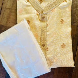 Pu&ograve; includere: Un completo kurta e pigiama color crema. La kurta presenta un colletto alla coreana e ricami dorati intricati lungo lo scollo e la paramontura. Il tessuto ha un sottile motivo floreale in pesca e oro. I pantaloni del pigiama sono color crema abbinato.