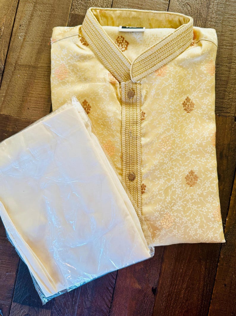 Pu&ograve; includere: Un completo kurta e pigiama color crema. La kurta presenta un colletto alla coreana e ricami dorati intricati lungo lo scollo e la paramontura. Il tessuto ha un sottile motivo floreale in pesca e oro. I pantaloni del pigiama sono color crema abbinato.