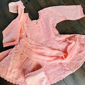 May include: A light pink embroidered outfit, including a long-sleeved top, pants, and a scarf. The top features a round neckline and decorative buttons. The fabric has a delicate, openwork pattern. The outfit is displayed on a dark wooden surface.