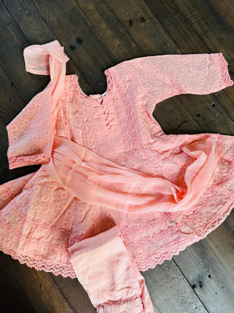 May include: A light pink embroidered outfit, including a long-sleeved top, pants, and a scarf. The top features a round neckline and decorative buttons. The fabric has a delicate, openwork pattern. The outfit is displayed on a dark wooden surface.