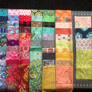 May include: A collection of fabric squares in various colors and patterns, including floral, geometric, and abstract designs. The squares are arranged in rows and columns on a gray surface.