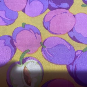 May include: Close-up of a fabric featuring a repeating pattern of purple and pink plums on a yellow backdrop. Some plums are whole, while others are cut open, revealing a brown stone. The plums have green stalks.