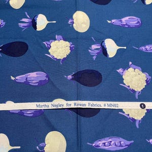 May include: Dark blue fabric featuring a repeating pattern of stylised vegetables in white, purple, and cream. The design includes aubergines, cauliflower, and other produce. The text "Martha Negley for Rowan Fabrics. #MN02" is printed on a white strip.
