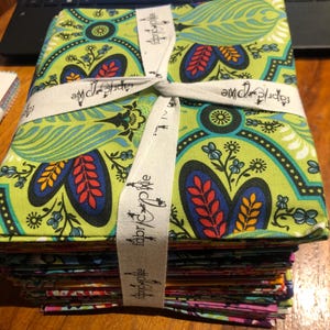 Könnte beinhalten: Ein Stapel gemusterter Stoffquadrate, gebunden mit einem weißen Band. Der Stoff hat einen grünen Hintergrund mit blauen, roten und gelben Blumenmustern. Auf dem Band steht "fabric pixie".