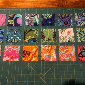 Peut inclure: Une collection de 16 carrés de tissu, chacun avec un motif coloré différent. Les carrés sont disposés en grille sur un tapis de coupe vert. Les motifs comprennent des motifs floraux, géométriques et abstraits.