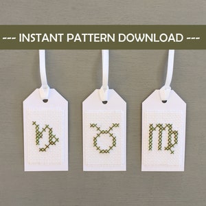 May include: Three white tags with cross-stitch zodiac symbols: Capricorn, Taurus, and Virgo. Each tag has a white ribbon for hanging. The text "--- INSTANT PATTERN DOWNLOAD ---" is above the tags.