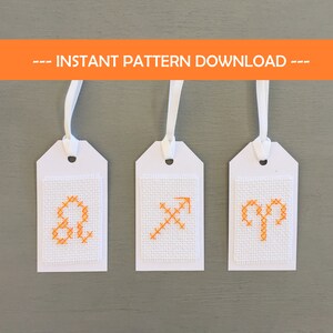Könnte beinhalten: Drei weiße Anhänger mit orangefarbenen Kreuzstich-Tierkreiszeichen: Löwe, Schütze und Widder. Jeder Anhänger hat ein weißes Band und den Text "-- INSTANT PATTERN DOWNLOAD --" in einem orangefarbenen Banner.
