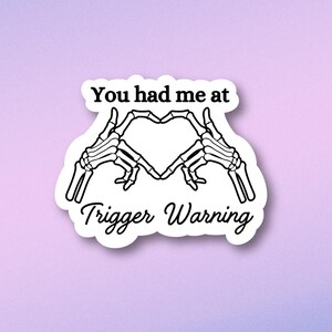 May include: A sticker with a skeleton hand making a heart shape with the text "You had me at Trigger Warning".