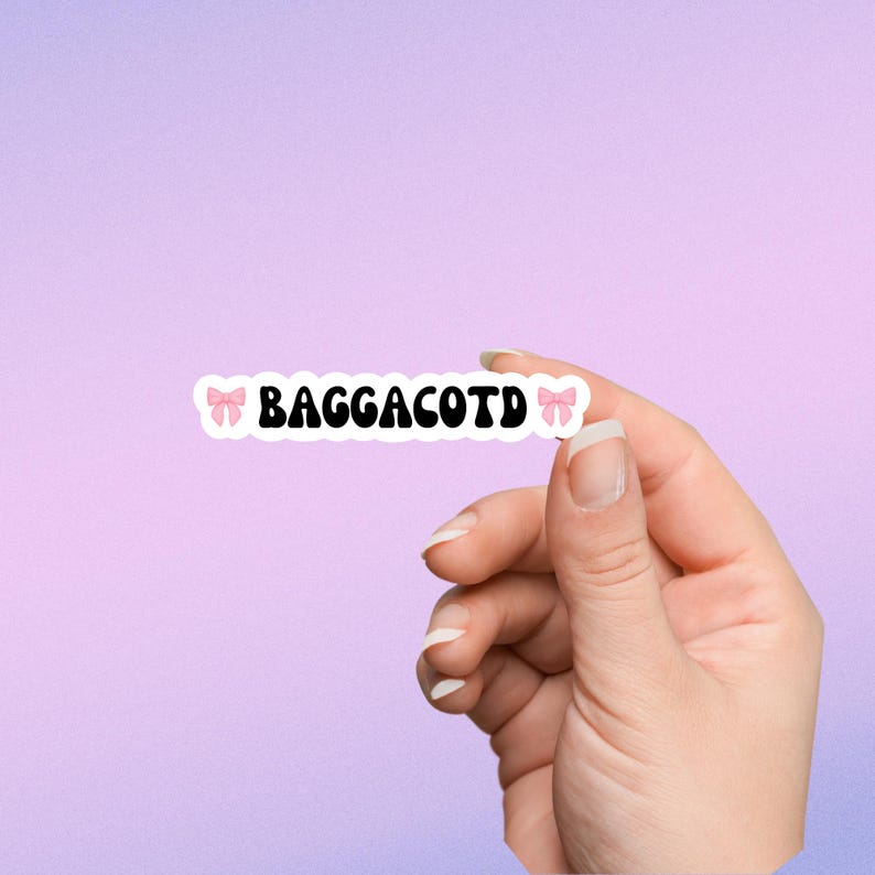 Op de afbeelding: Een witte sticker met roze strikken aan beide kanten en de tekst "BAGGACOTD" in zwart.