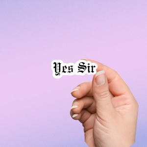 May include: A white sticker with black text that reads "Yes Sir" in a gothic font. A hand is holding the sticker.