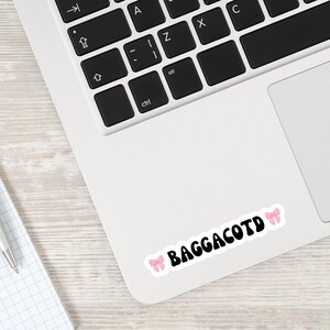Op de afbeelding: Een laptop met een sticker waarop "Baggacotd" staat, met twee roze strikken aan weerszijden van de tekst.