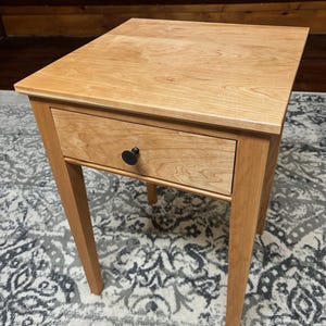 May include: A light-colored wooden side table with a single drawer and a black heart-shaped knob. The table has a rectangular top and four straight legs. The drawer is centered below the tabletop. The table is approximately 24 inches tall.