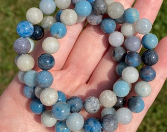 May include: A bracelet made of blue, gray, and white gemstone beads. The beads are round and have a smooth, polished finish.