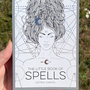 May include: A grey book with a silver foil design on the cover. The design features a woman with her eyes closed and a crescent moon on her forehead. The book title is "The Little Book of Spells" by Astrid Carvel.