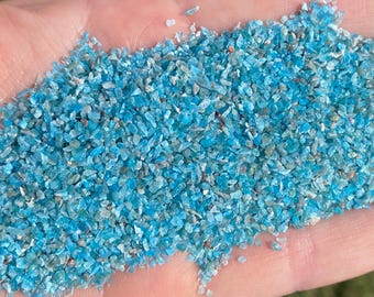 May include: A close-up of a handful of small, light blue, iridescent glass chips. The chips are irregularly shaped and have a sparkly, glittery appearance.