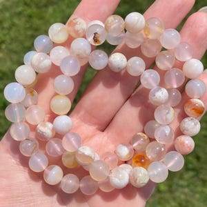Puede incluir: Un brazalete hecho de cuentas de ágata blanca con inclusiones marrones y naranjas. Las cuentas son redondas y lisas, y el brazalete está enhebrado en un cordón elástico transparente.