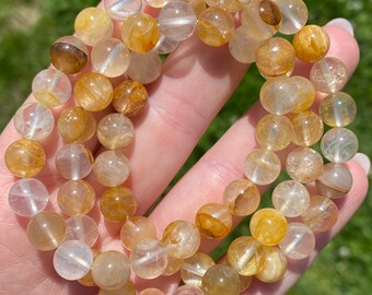 May include: A gold rutilated quartz bracelet with clear and golden beads. The bracelet is made of natural stones and is approximately 8 inches in length.