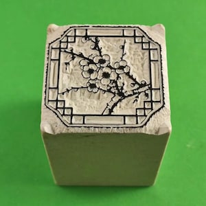 Puede incluir: Un sello cuadrado blanquecino con un diseño floral negro. El diseño presenta una rama con flores dentro de un borde rectangular. El sello está sobre un fondo verde.