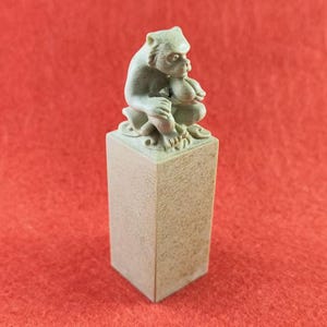 Puede incluir: Una escultura de piedra tallada de un mono sentado sobre una base rectangular. El mono es de color gris claro y parece estar sosteniendo algo. La base es de color beige claro. El fondo es rojo liso.