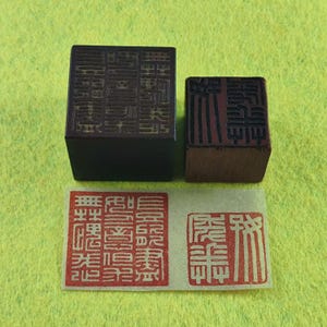 Puede incluir: Dos sellos chinos cuadrados de color marrón oscuro con intrincadas tallas. Debajo hay un papel rectangular con dos impresiones de tinta roja de los sellos. Los sellos y las impresiones están sobre una superficie verde claro.