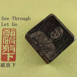Op de afbeelding: Een donkere, vierkante stempel met een gebeeldhouwde afbeelding van een Boeddha-figuur. De stempel is schuin geplaatst en toont het getextureerde oppervlak. Er staat ook tekst in het Engels en Chinese karakters.