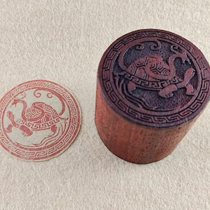 Puede incluir: Un sello de madera con un diseño de tortuga. El sello cilíndrico tiene un tallado detallado de tortuga en la parte superior. También hay una impresión circular del sello, que muestra el diseño de la tortuga en rojo.