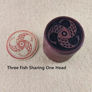 Puede incluir: Un sello redondo rojo y crema y un sello cilíndrico marrón oscuro con un diseño de pez. El texto "Three Fish Sharing One Head" está debajo de los sellos. Los sellos están sobre una superficie marrón claro.