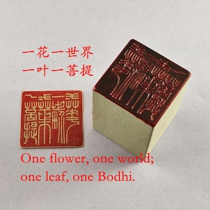 Puede incluir: Un sello de tinta roja con un diseño detallado y una almohadilla rectangular. El texto "Una flor, un mundo; una hoja, un Bodhi" está impreso en rojo. El sello se utiliza probablemente para arte o caligrafía.