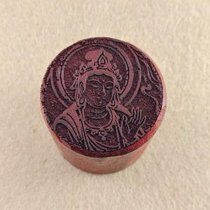 Puede incluir: Un sello cilíndrico de color marrón rojizo con una imagen tallada de una figura serena, posiblemente una deidad, rodeada por un halo. La figura está detallada, con una corona y joyas ornamentadas. La superficie del sello tiene un aspecto ligeramente texturizado.