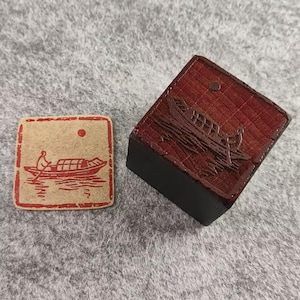 Puede incluir: Un sello de madera rojo y marrón y una imagen estampada. El sello presenta un barco con una persona remando. La imagen estampada está en un cuadrado beige con un barco rojo y un sol rojo. El sello es un cuadrado rojo oscuro con un diseño de barco.