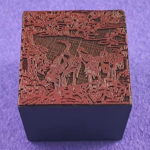 Puede incluir: Un bloque cuadrado azul oscuro con una parte superior marrón rojiza detallada. La parte superior presenta un diseño en relieve de grullas y árboles, creando una apariencia texturizada y artística. El conjunto sugiere un objeto decorativo o de colección.
