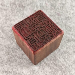 Puede incluir: Un sello cuadrado de madera con un diseño detallado en tinta roja en su superficie superior. Los lados del sello son de color marrón natural, y el intrincado patrón sugiere un significado tradicional o cultural. El sello probablemente se utiliza para imprimir.