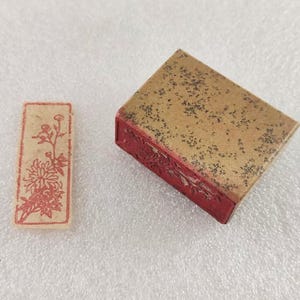 Puede incluir: Un sello rectangular y una caja de sellos cuadrada. El sello tiene un diseño floral rojo sobre un fondo beige. La caja del sello es marrón con motas negras y lados rojos. Los artículos son probablemente para arte o artesanía.
